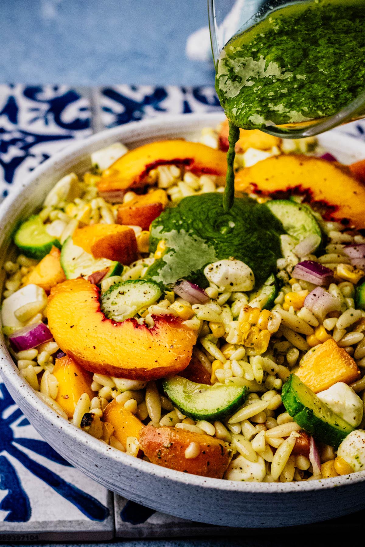 Pouring basil vinaigrette over summer orzo salad with grilled peaches, corn, cucumber, mozzarella, and red onion.