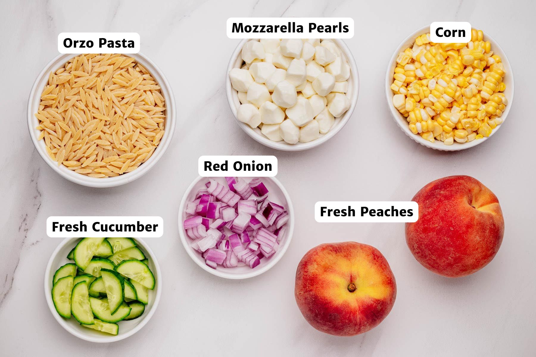 Labeled ingredients for summer orzo salad on a white surface, including orzo pasta, mozzarella pearls, corn, red onion, cucumber, and fresh peaches.