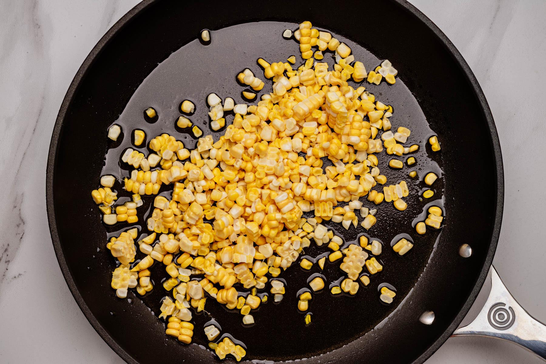Corn kernels sautéing in olive oil in a black skillet on a white surface.