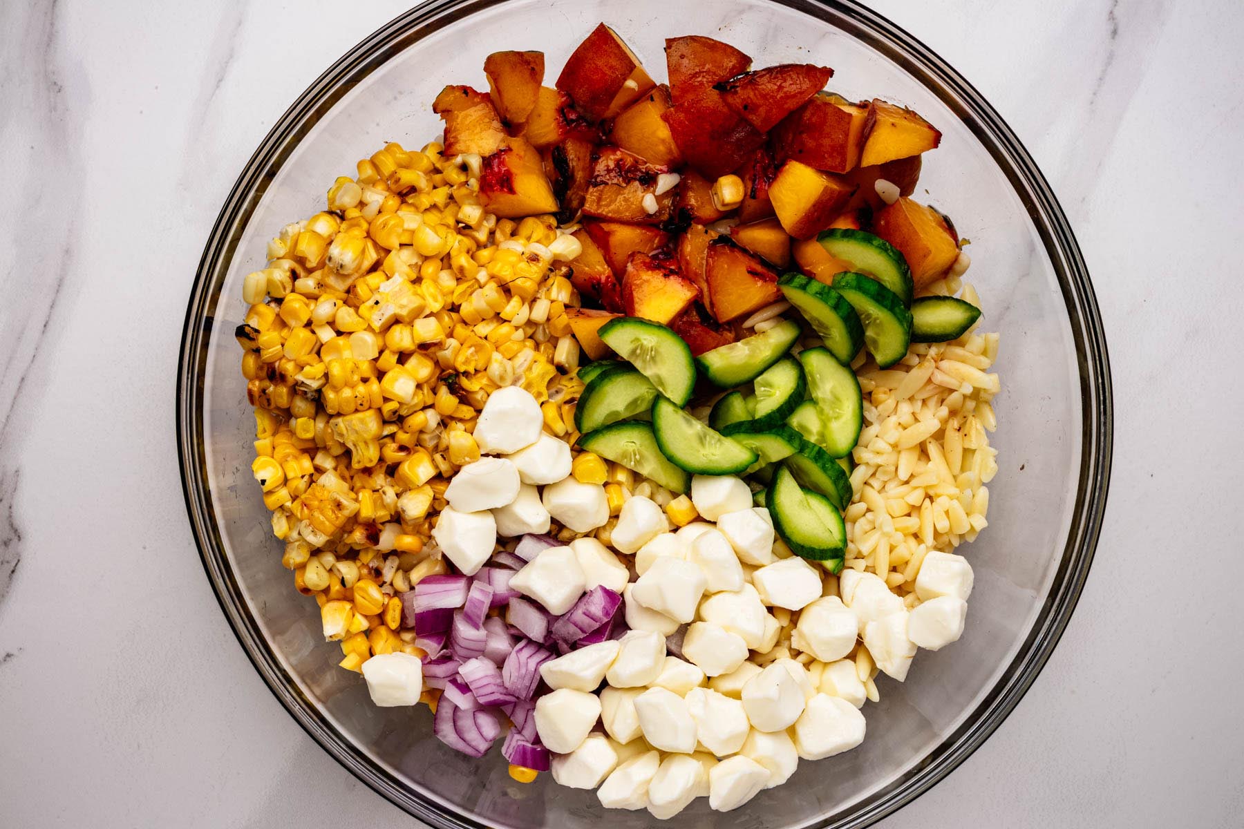 Grilled corn, peaches, cucumber, red onion, mozzarella, and orzo arranged in sections in a glass mixing bowl before tossing.