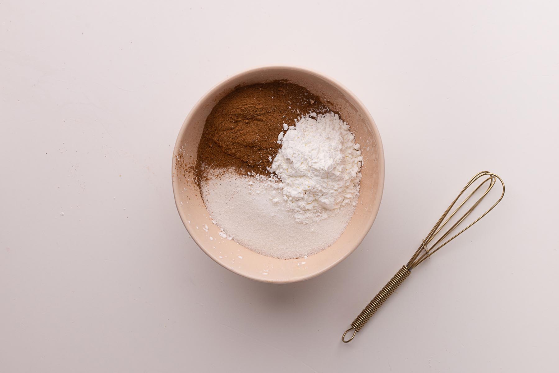 Small bowl with unmixed cinnamon, cornstarch, and granulated sugar sitting on a light surface next to a small gold whisk.