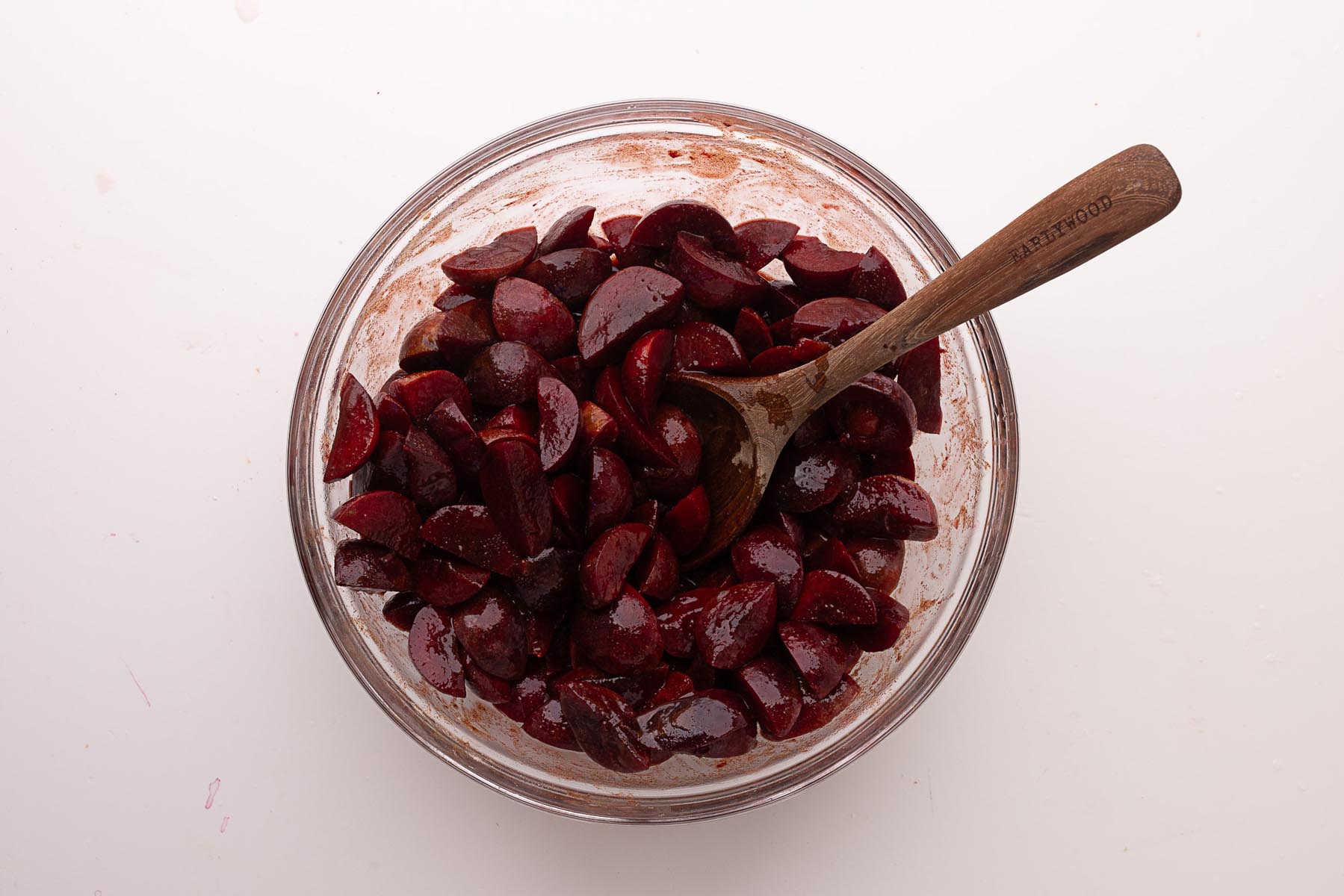 Quartered plums in a glass bowl being stirred with a wooden spoon after adding bourbon and lemon juice.