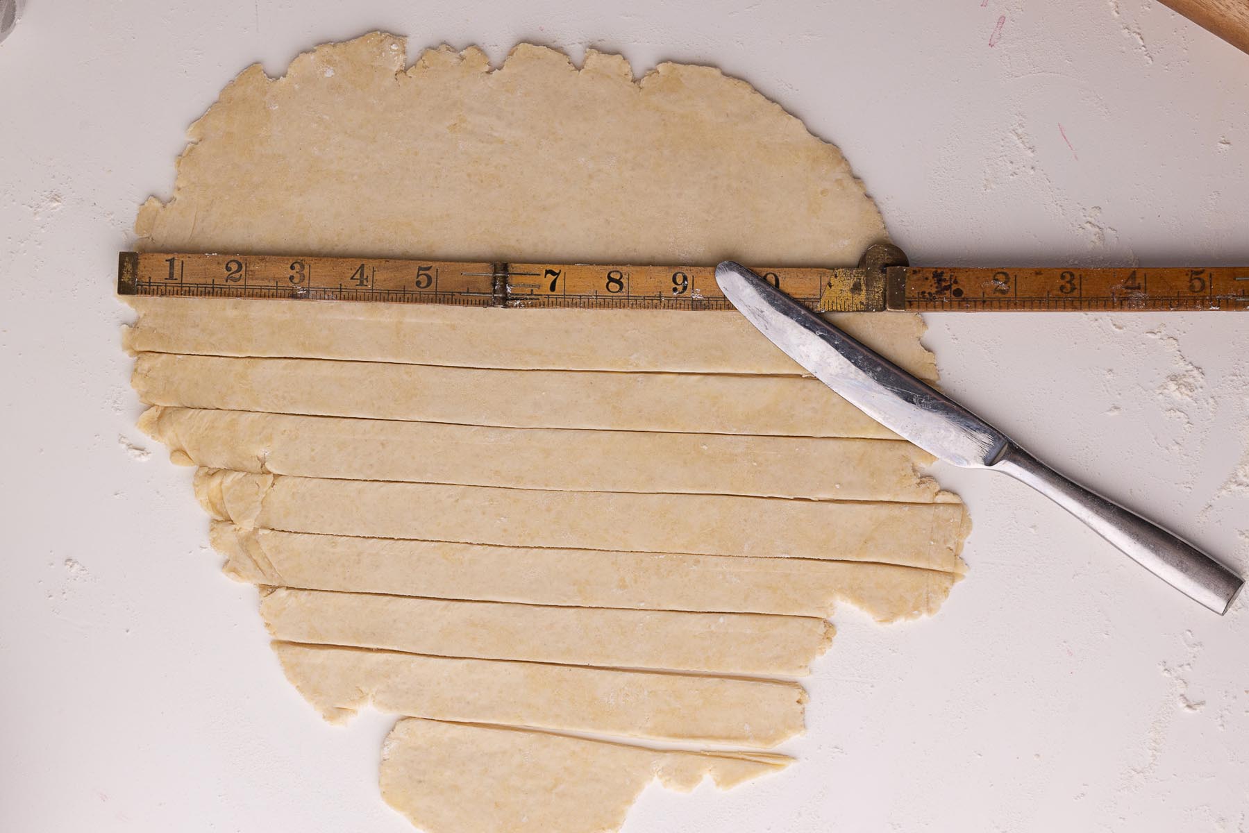 Rolled-out pie dough cut into strips using a knife and a wooden ruler for measuring, set on a floured white surface.