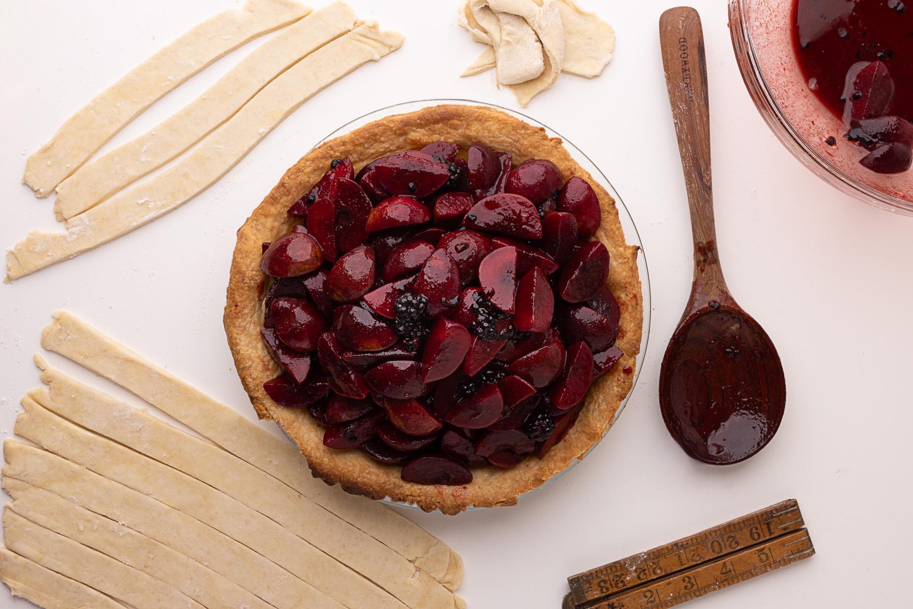 Par-baked pie crust filled with glossy plum and blackberry mixture, surrounded by strips of raw pie dough ready for lattice, a wooden spoon with filling residue, and a measuring stick on a white surface.