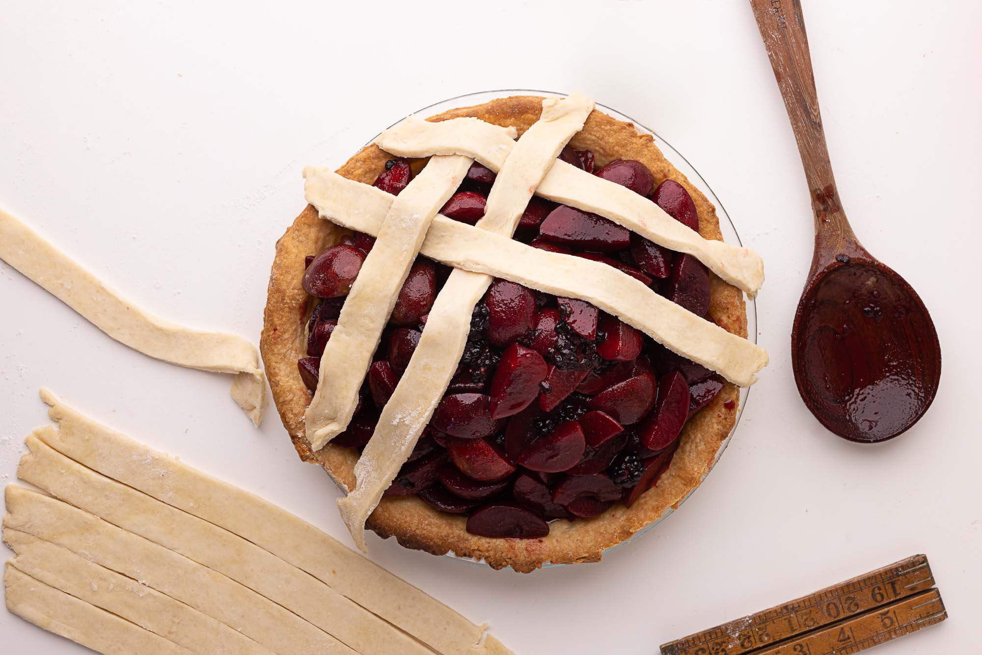 Partially assembled fruit pie with sliced plums and blueberries, topped with raw lattice dough strips. A wooden spoon with fruit juices, dough scraps, and a folding ruler rest nearby on a floured white surface.