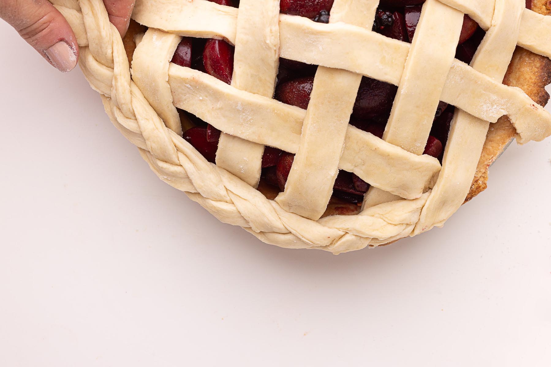 Close-up of a hand holding a pie with woven lattice crust and braided edge, filled with glossy plum and blackberry filling, ready for baking.
