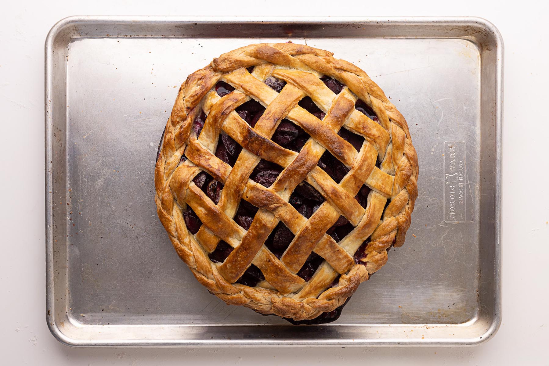 Fully baked plum and blackberry pie with a golden brown lattice crust and braided edge, resting on a metal baking sheet.