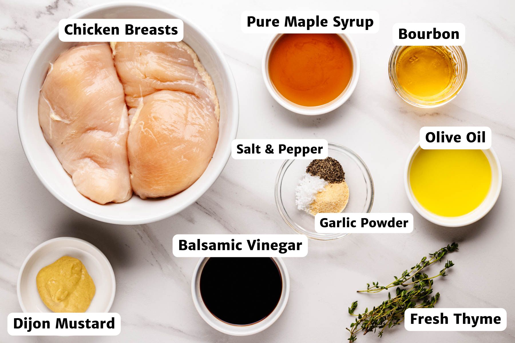 Overhead view of labeled ingredients for bourbon glazed chicken, including chicken breasts, pure maple syrup, bourbon, olive oil, Dijon mustard, balsamic vinegar, fresh thyme, salt, pepper, and garlic powder.