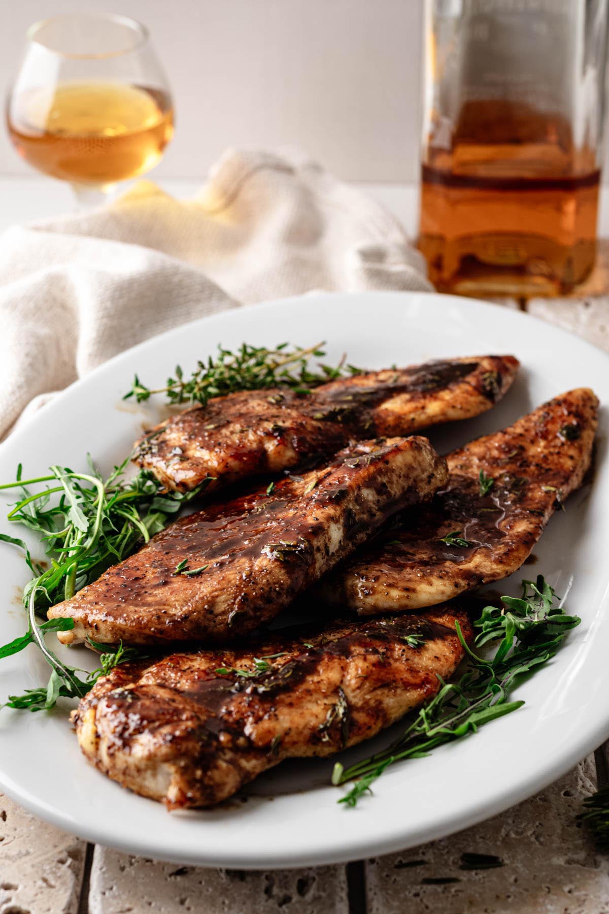 Golden-brown chicken cutlets drizzled with a glossy bourbon-maple glaze, garnished with fresh thyme, arranged on a white platter with a glass and bottle of bourbon in the background.