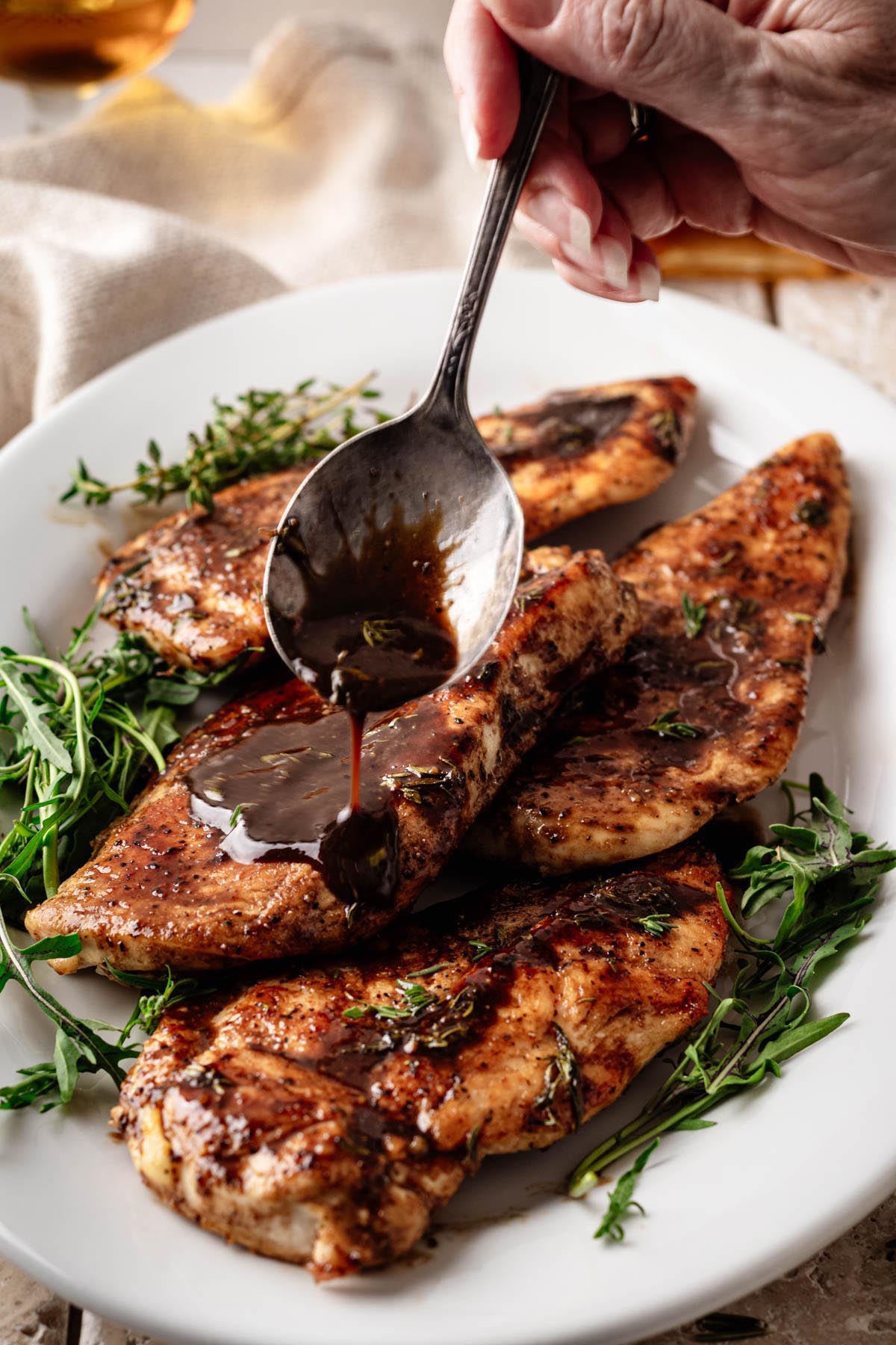 A hand spooning glossy bourbon-maple glaze over golden-brown chicken cutlets, garnished with fresh thyme, on a white platter with fresh herbs.