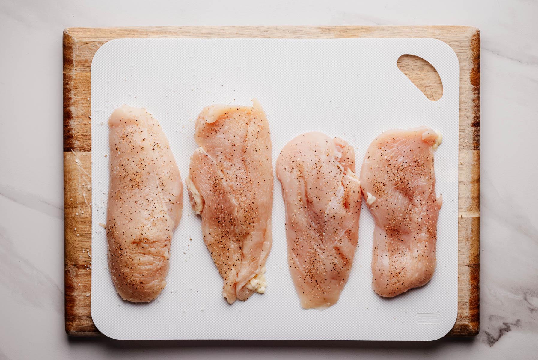 Raw chicken cutlets seasoned with salt and pepper, arranged in a row on a white cutting board set over a wooden board.