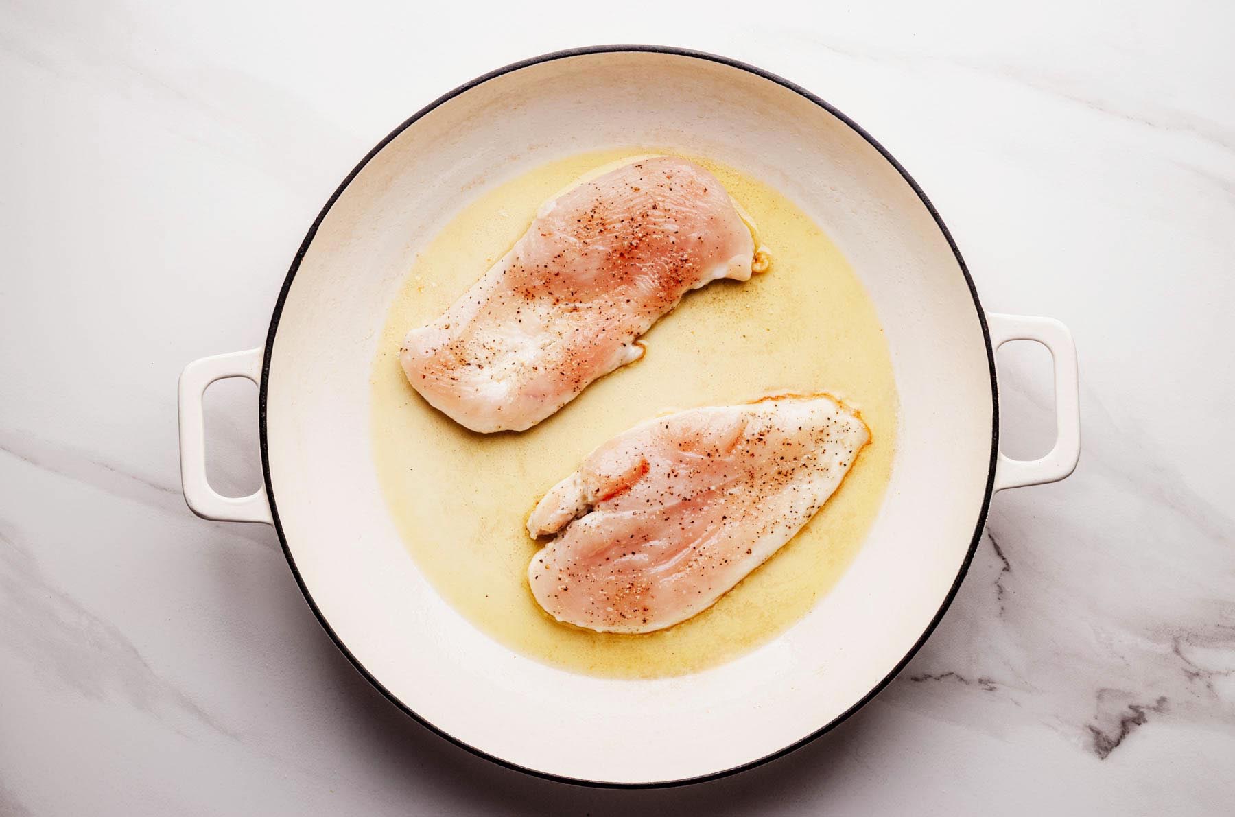 Seasoned chicken cutlets searing in olive oil in a white enameled skillet.