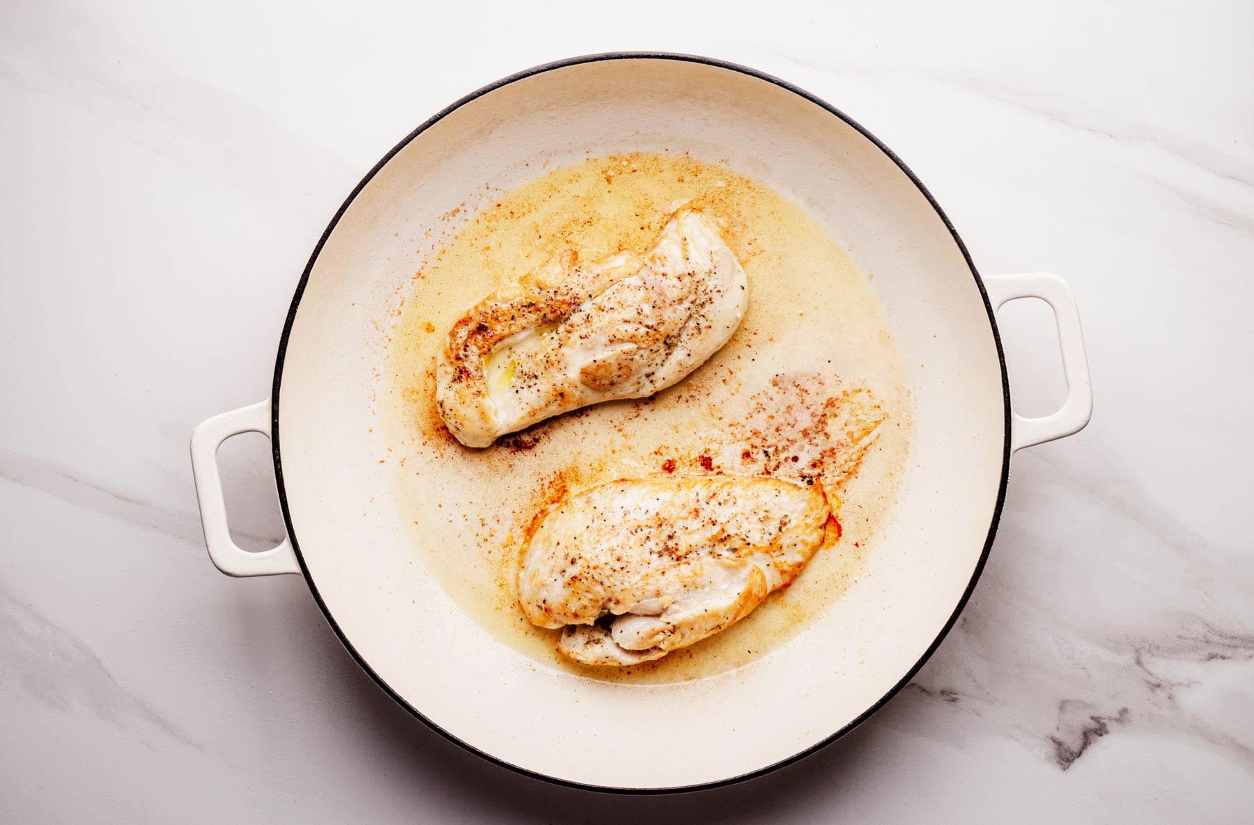 Partially seared chicken cutlets in a white enameled skillet, beginning to turn golden on the outside while cooking in olive oil.