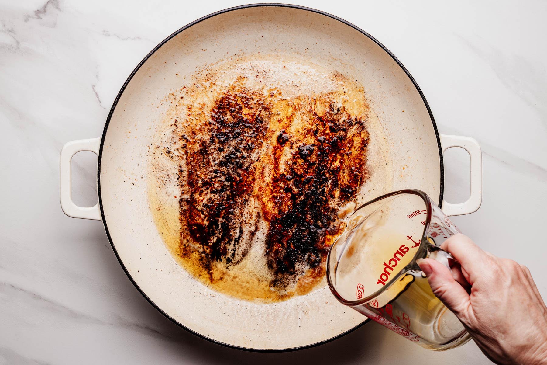 Bourbon being poured into a white enameled skillet with browned bits after searing chicken, ready to deglaze the pan and start the glaze.
