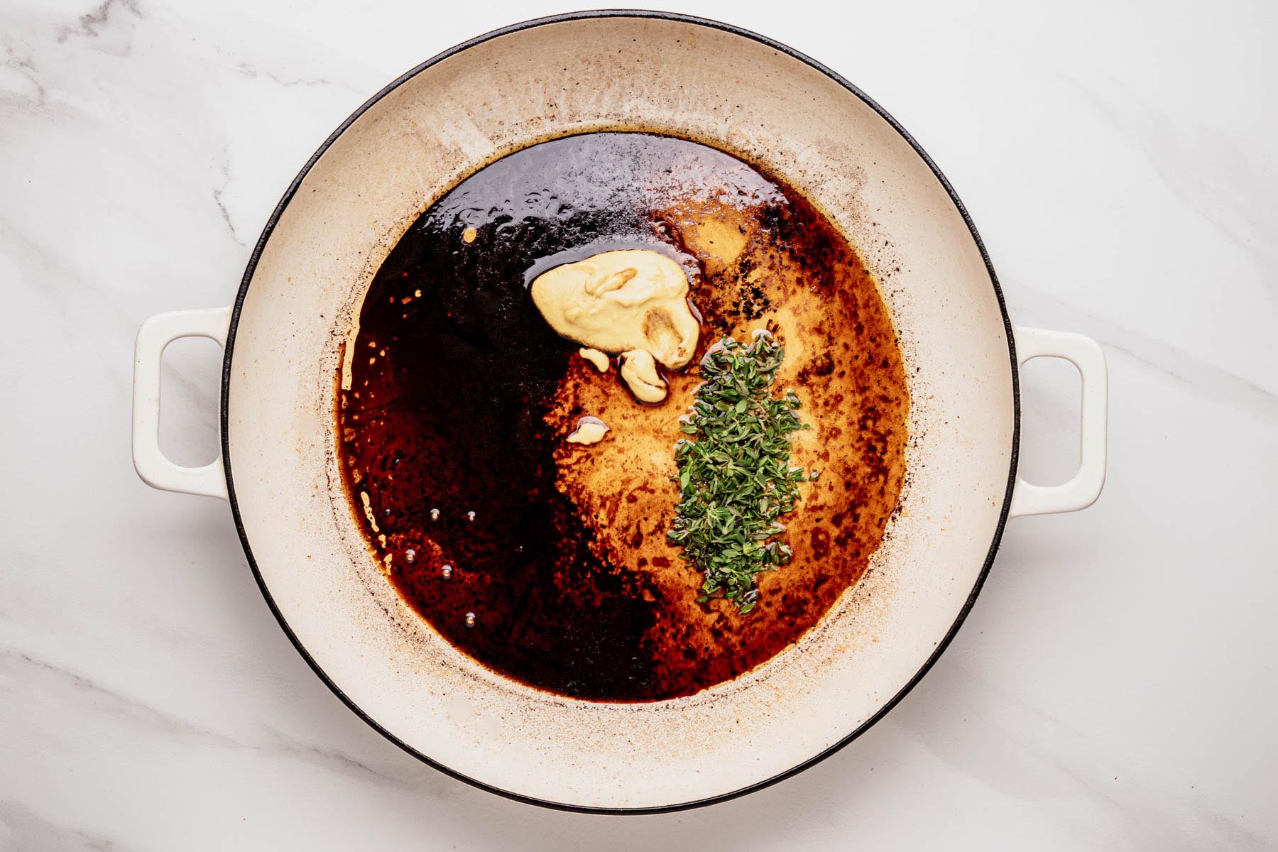 Bourbon-maple glaze ingredients &mdash; Dijon mustard, fresh thyme leaves, and balsamic mixture &mdash; in a white enameled skillet, ready to be whisked together.