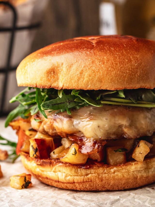 Juicy ground pork burger with melted cheese, saut&eacute;ed apples, and fresh arugula on a toasted brioche bun, with fries in the background.