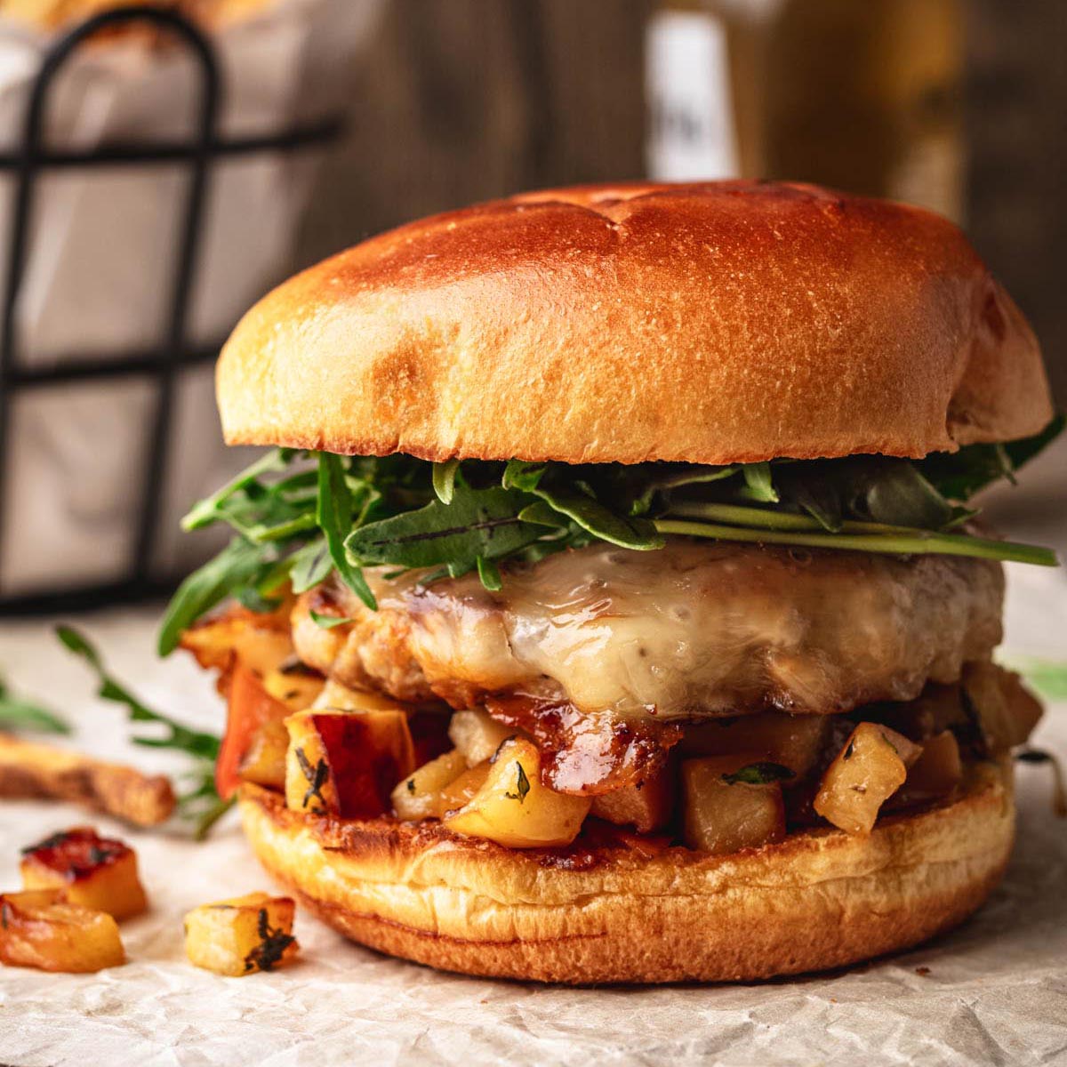 Juicy ground pork burger with melted cheese, sautéed apples, and fresh arugula on a toasted brioche bun, with fries in the background.