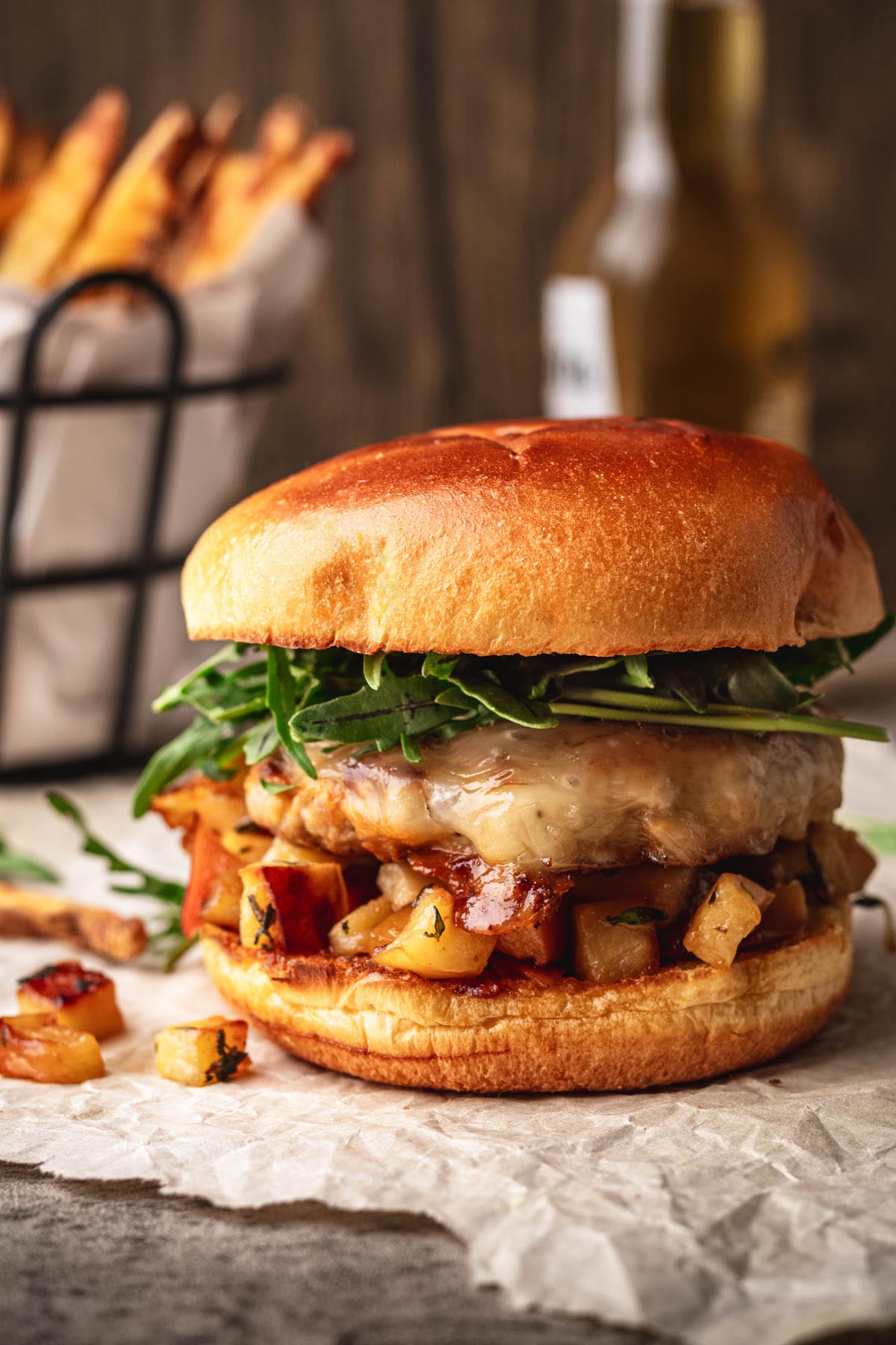 Juicy ground pork burger with melted cheese, saut&eacute;ed apples, and fresh arugula on a toasted brioche bun, with fries in the background.