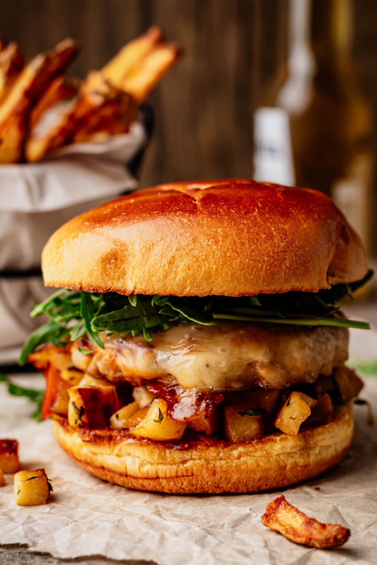 Juicy ground pork burger with melted cheese, saut&eacute;ed apples, and fresh arugula on a toasted brioche bun, with fries in the background.