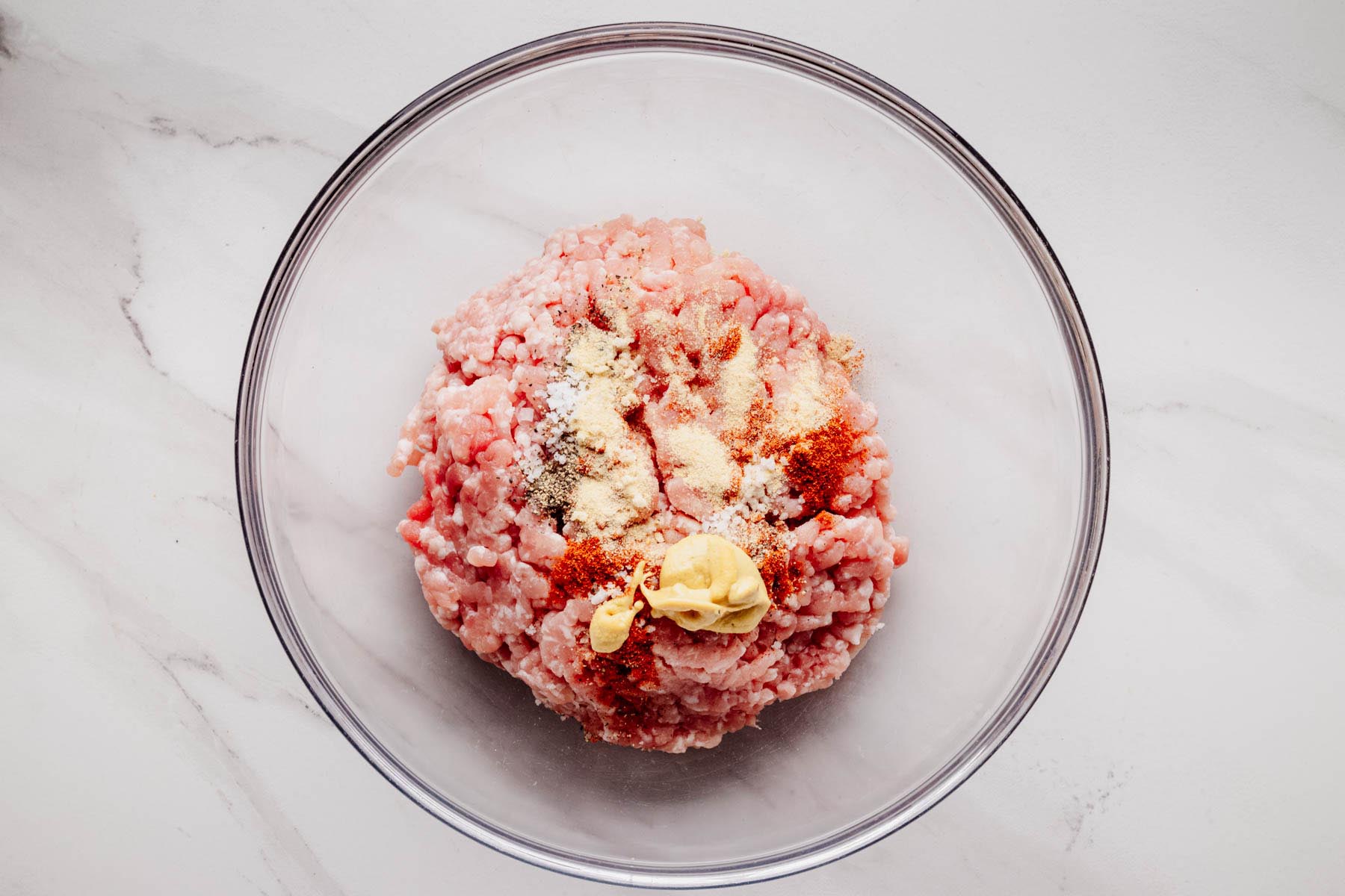 Ground pork in a mixing bowl topped with seasonings, smoked paprika, and Dijon mustard, ready to be combined for burger patties.