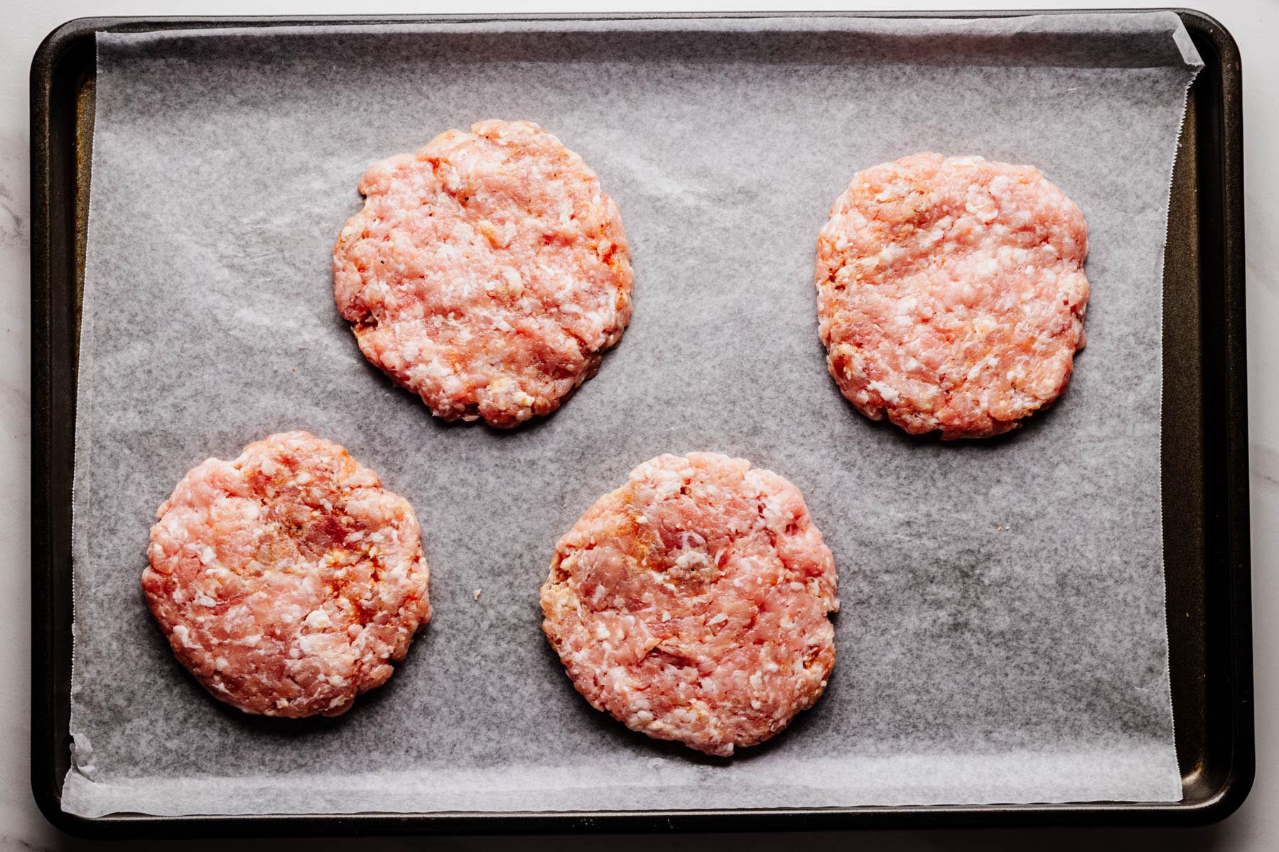 Four uncooked ground pork burger patties arranged on a parchment-lined baking sheet.