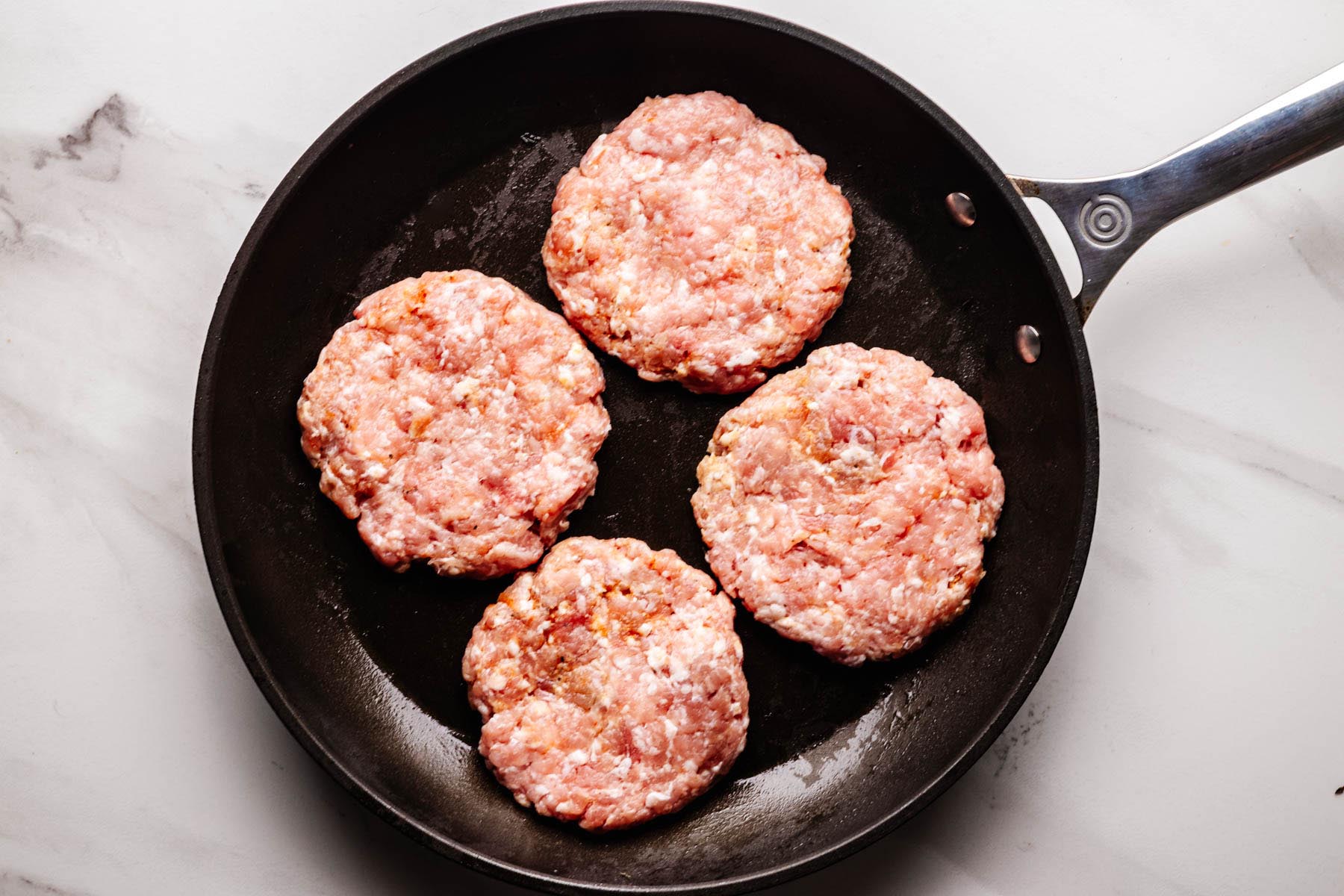 Four uncooked ground pork burger patties in a skillet, ready to be cooked.
