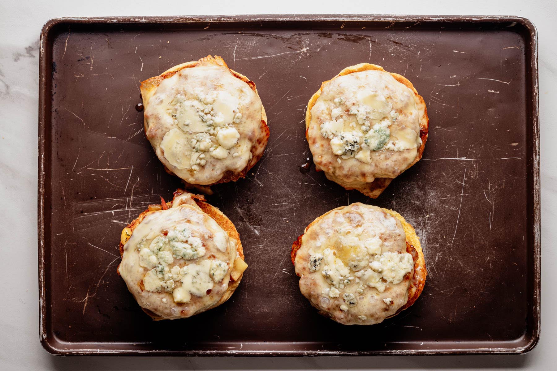 Toasted burger buns topped with saut&eacute;ed apples, ground pork patties, melted Muenster cheese, and crumbled Gorgonzola, arranged on a baking sheet.