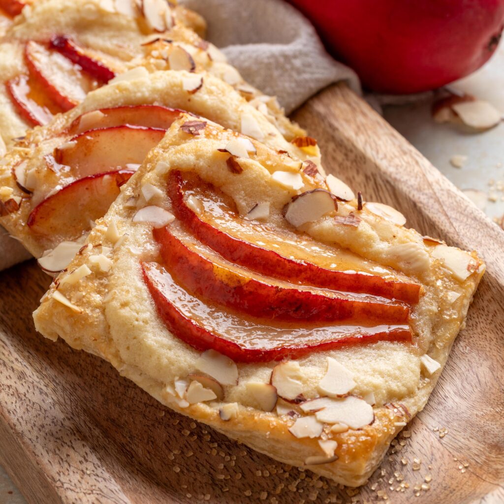 Close-up of a baked pear puff pastry tart topped with red pear slices and slivered almonds, displayed on a wooden board with coarse sugar scattered around.