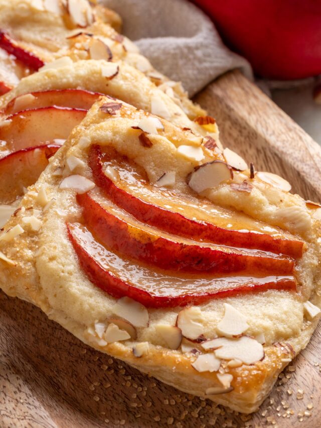 Close-up of a baked pear puff pastry tart topped with red pear slices and slivered almonds, displayed on a wooden board with coarse sugar scattered around.