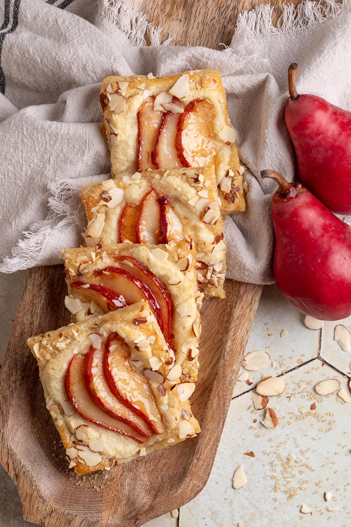 Golden brown pear puff pastry tarts topped with sliced red pears and slivered almonds, arranged on a wooden board with two fresh red pears on the side.