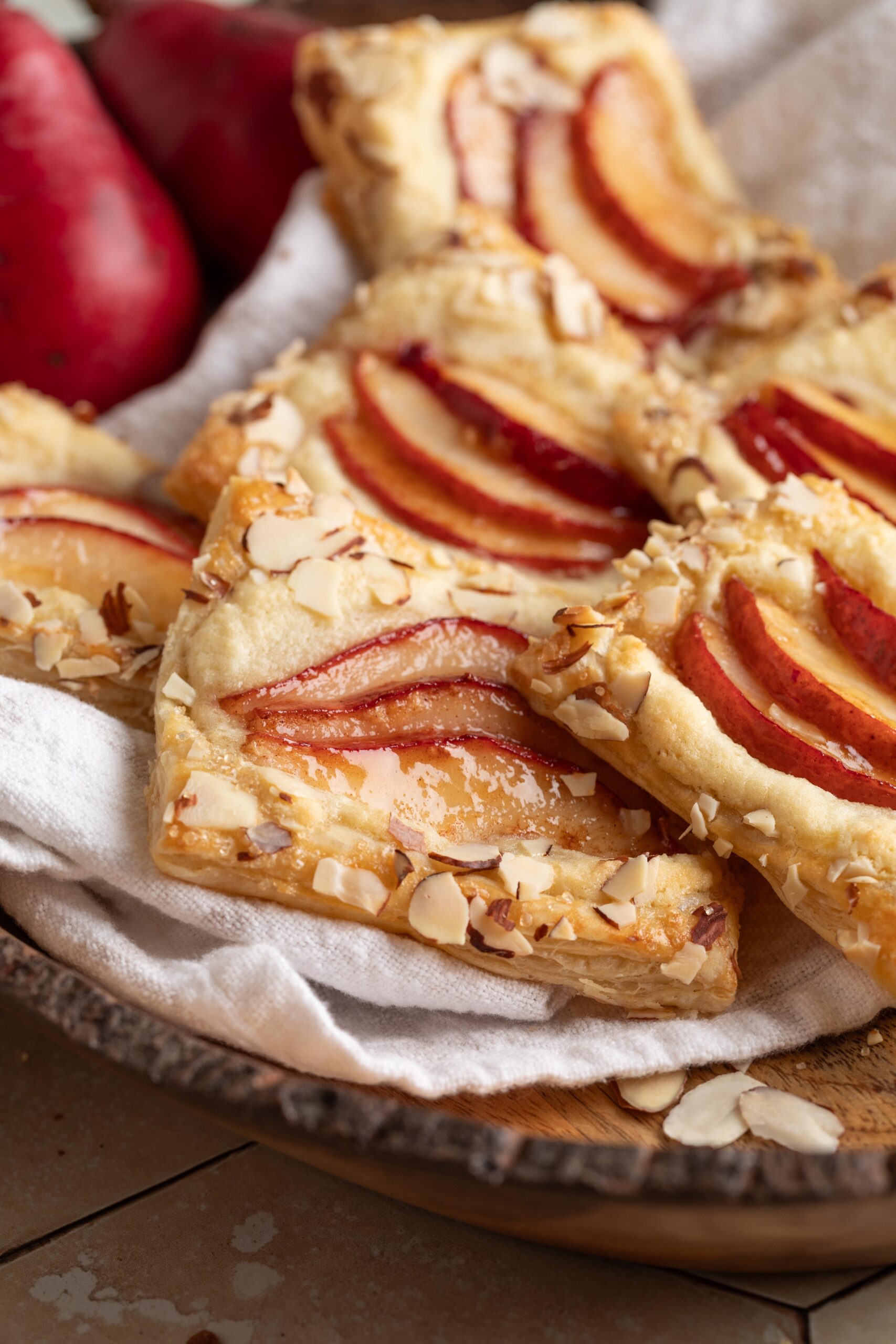 Pear puff pastry tarts topped with red pear slices and slivered almonds, arranged on a linen-lined wooden plate with fresh red pears in the background.