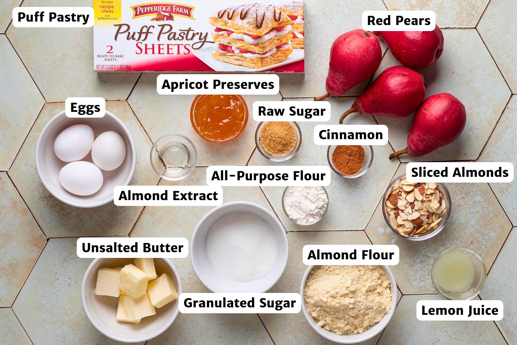 Overhead view of labeled ingredients for pear puff pastry tarts, including puff pastry sheets, red pears, eggs, apricot preserves, almond flour, sliced almonds, cinnamon, lemon juice, almond extract, all-purpose flour, granulated sugar, raw sugar, and unsalted butter.