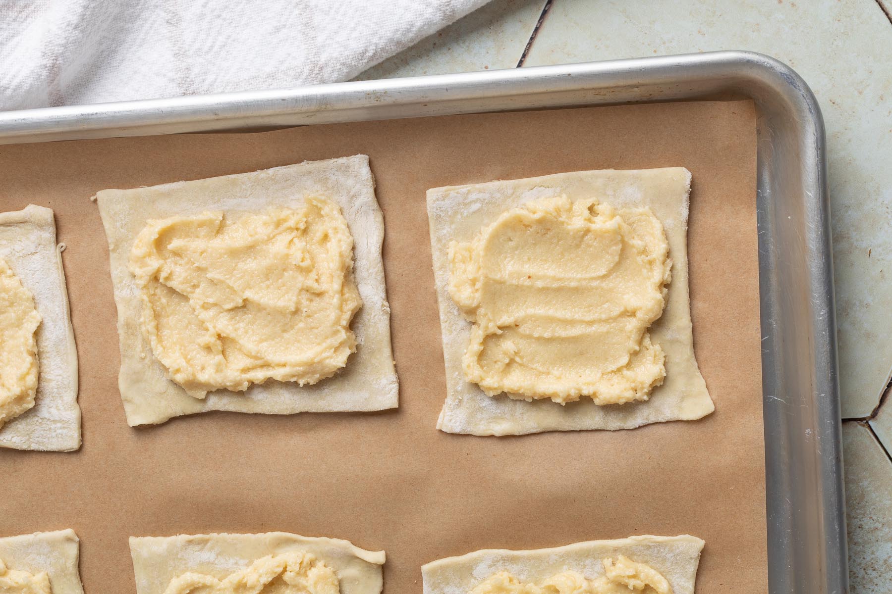 Squares of puff pastry on a parchment-lined baking sheet, each topped with a generous layer of almond cream spread evenly in the center, ready for sliced pears.