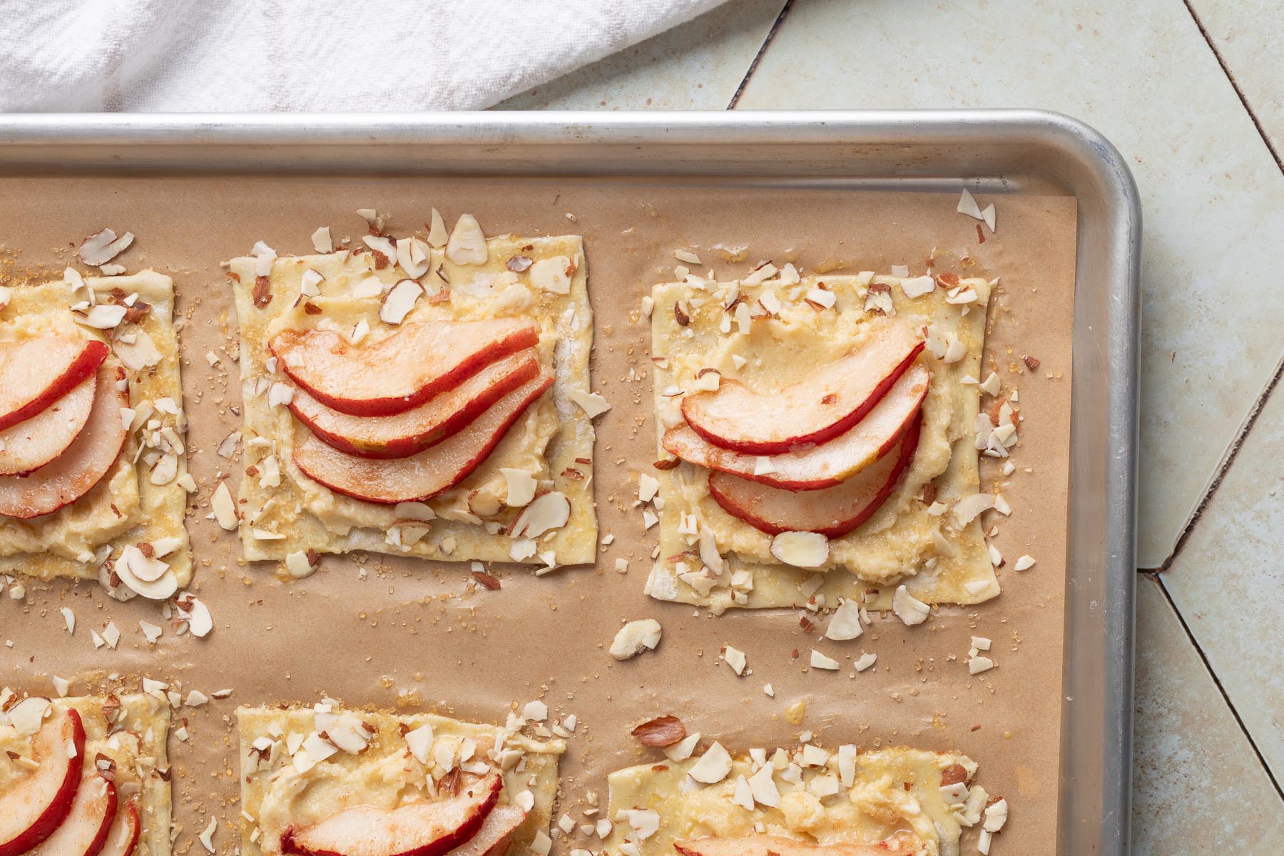 Unbaked pear puff pastry tarts on a parchment-lined baking sheet, each topped with almond cream, fanned red pear slices, sliced almonds, and coarse sugar, ready to go into the oven.