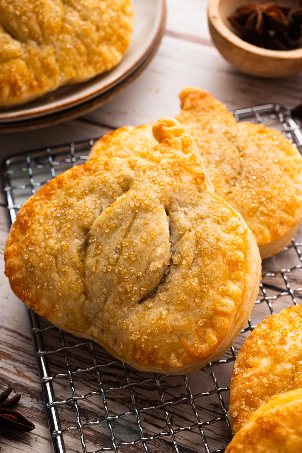 Close-up of golden brown pumpkin pie hand pies with coarse sugar topping, cooling on a wire rack with star anise and rustic plates in the background.