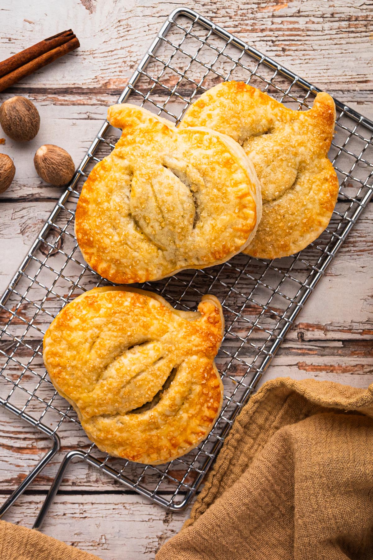 Mini pumpkin hand pies cooling on a rack, flaky crust with coarse sugar, styled with warm fall spices on a rustic wood background.