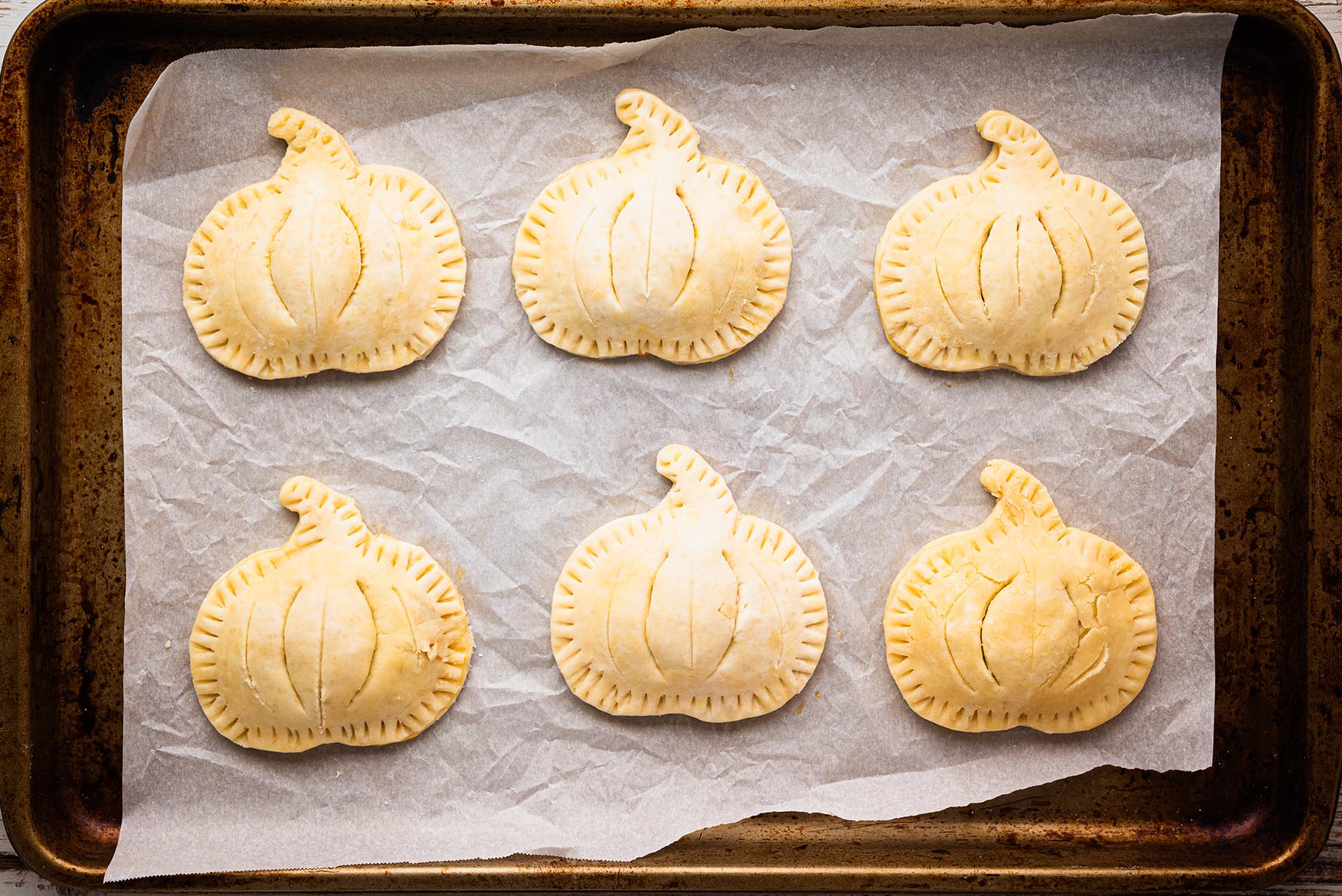Unbaked pumpkin-shaped hand pies on a parchment-lined baking sheet, edges crimped with a fork and decorative steam vents cut into the tops.
