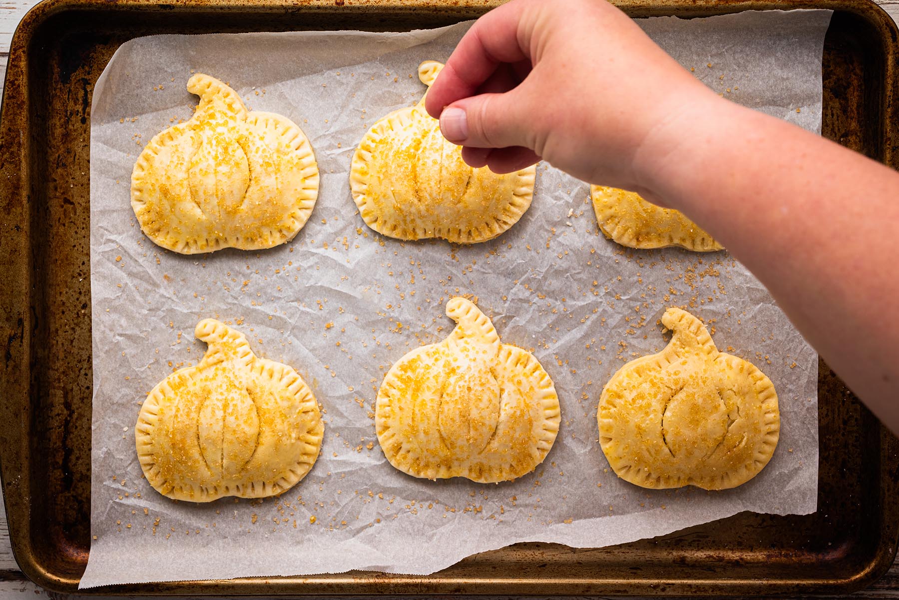 Unbaked pumpkin-shaped hand pies on a parchment-lined baking sheet, edges crimped and brushed with egg wash, while a hand sprinkles coarse sugar on top before baking.
