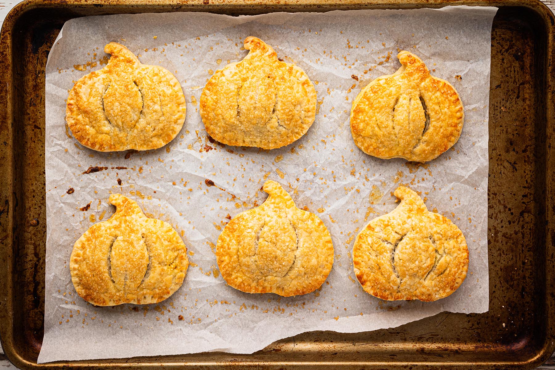 Freshly baked pumpkin-shaped hand pies on a parchment-lined baking sheet, golden brown with sparkling coarse sugar on top.