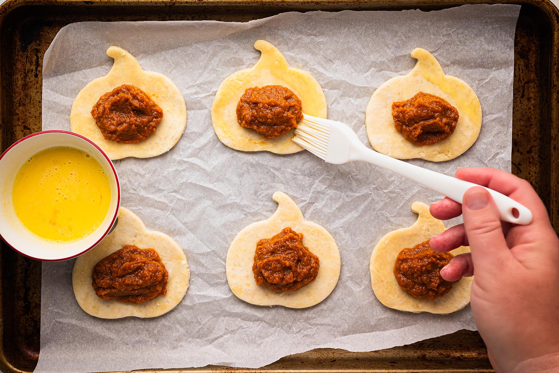 Pumpkin-shaped pie dough rounds topped with spiced pumpkin filling on a parchment-lined baking sheet, with a hand brushing egg wash around the edges to prepare for sealing.