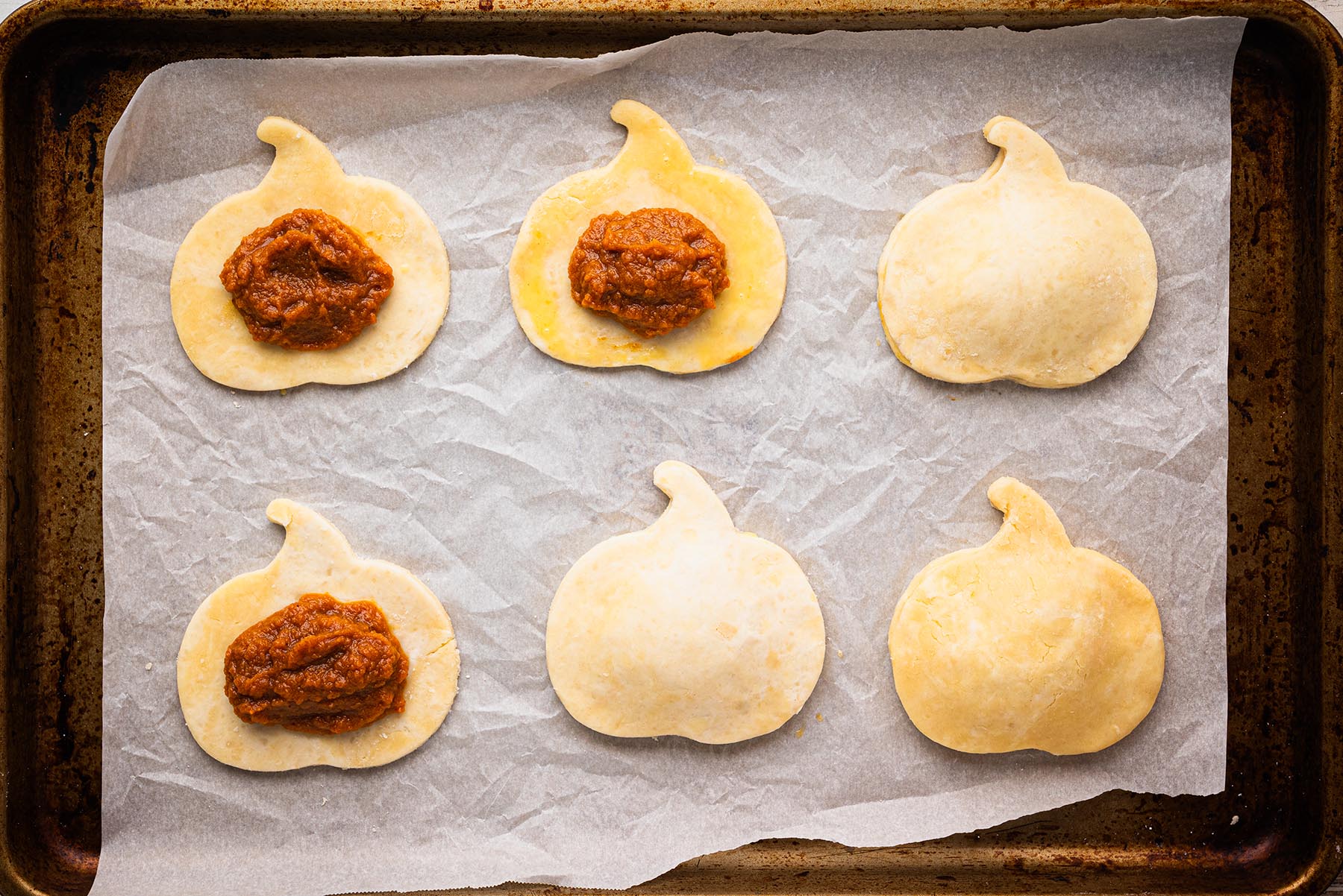 Pumpkin-shaped pie dough cutouts on a baking sheet, some filled with pumpkin filling and others topped with a second layer of dough, ready to be sealed into hand pies.