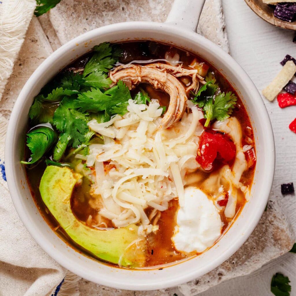 Bowl of Southwestern chicken soup topped with shredded cheese, avocado slices, cilantro, and sour cream.