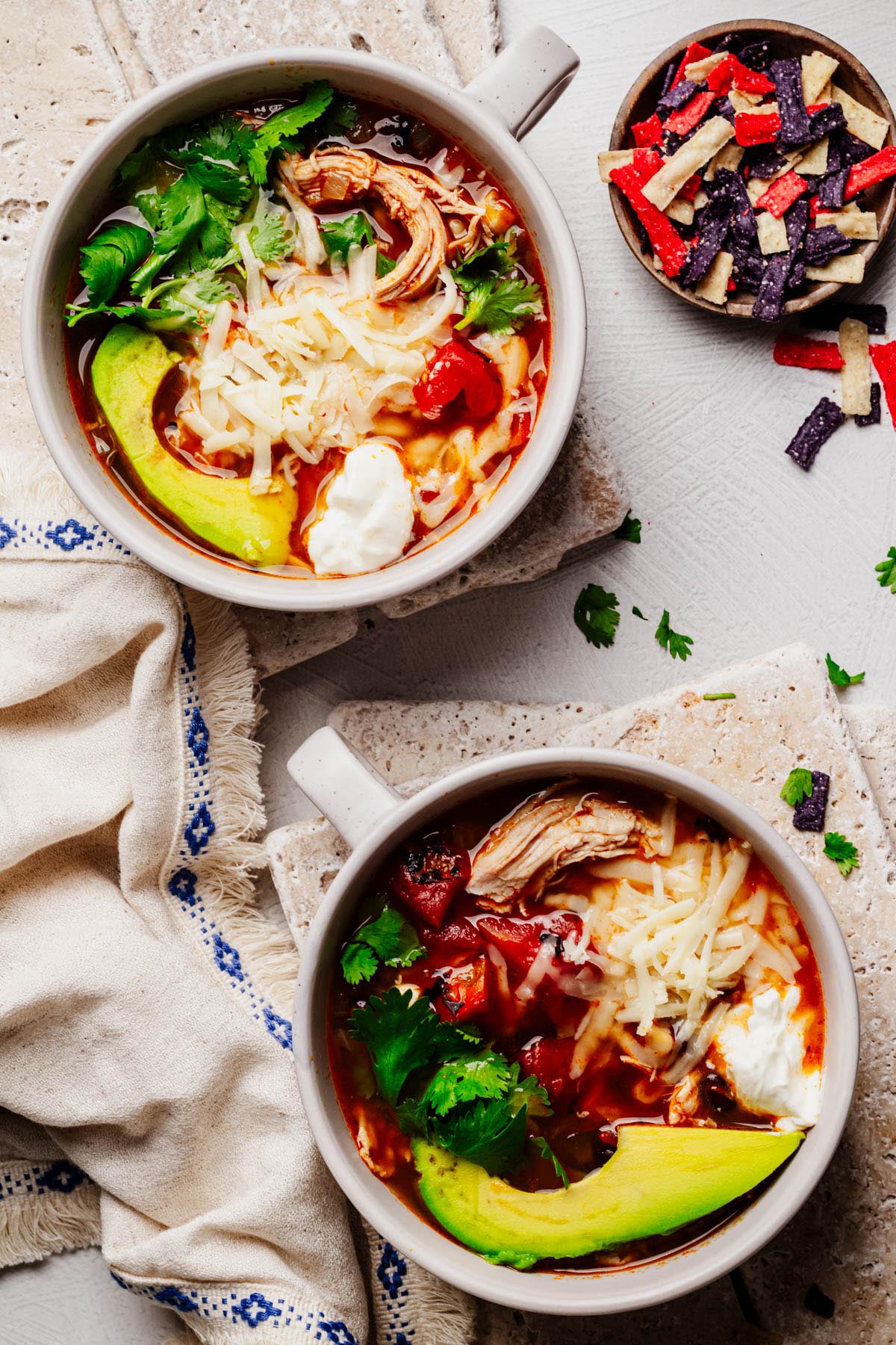 Two bowls of Southwestern chicken soup topped with shredded cheese, avocado slices, cilantro, and sour cream, served with tortilla strips on the side.