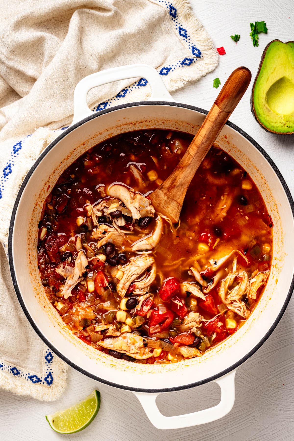 White Dutch oven filled with Southwestern chicken soup with shredded chicken, black beans, corn, and tomatoes, stirred with a wooden spoon.