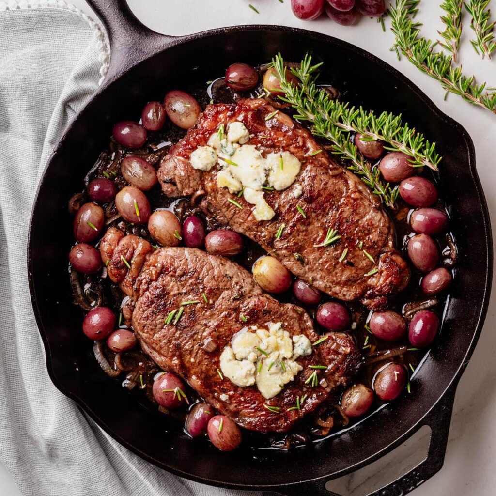 Two seared strip steaks in a cast iron skillet, topped with blue cheese and garnished with rosemary, surrounded by roasted red grapes and caramelized shallots.