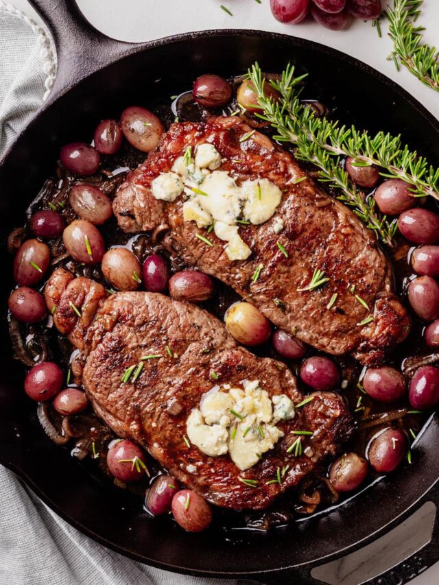 Two seared strip steaks in a cast iron skillet, topped with blue cheese and garnished with rosemary, surrounded by roasted red grapes and caramelized shallots.