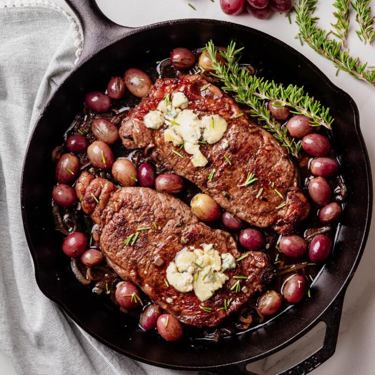 Two seared strip steaks in a cast iron skillet, topped with blue cheese and garnished with rosemary, surrounded by roasted red grapes and caramelized shallots.