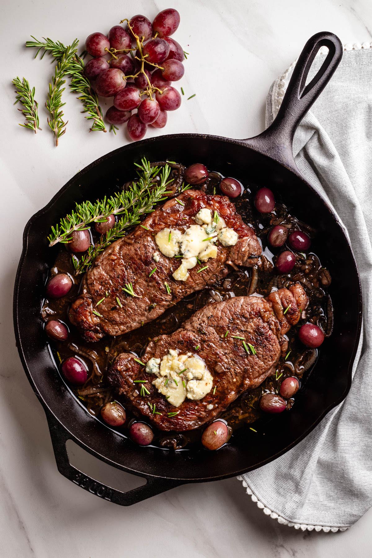 Cast iron skillet with two skillet steaks topped with blue cheese, roasted grapes, caramelized shallots, and fresh rosemary sprigs, styled with grapes and herbs on the side.