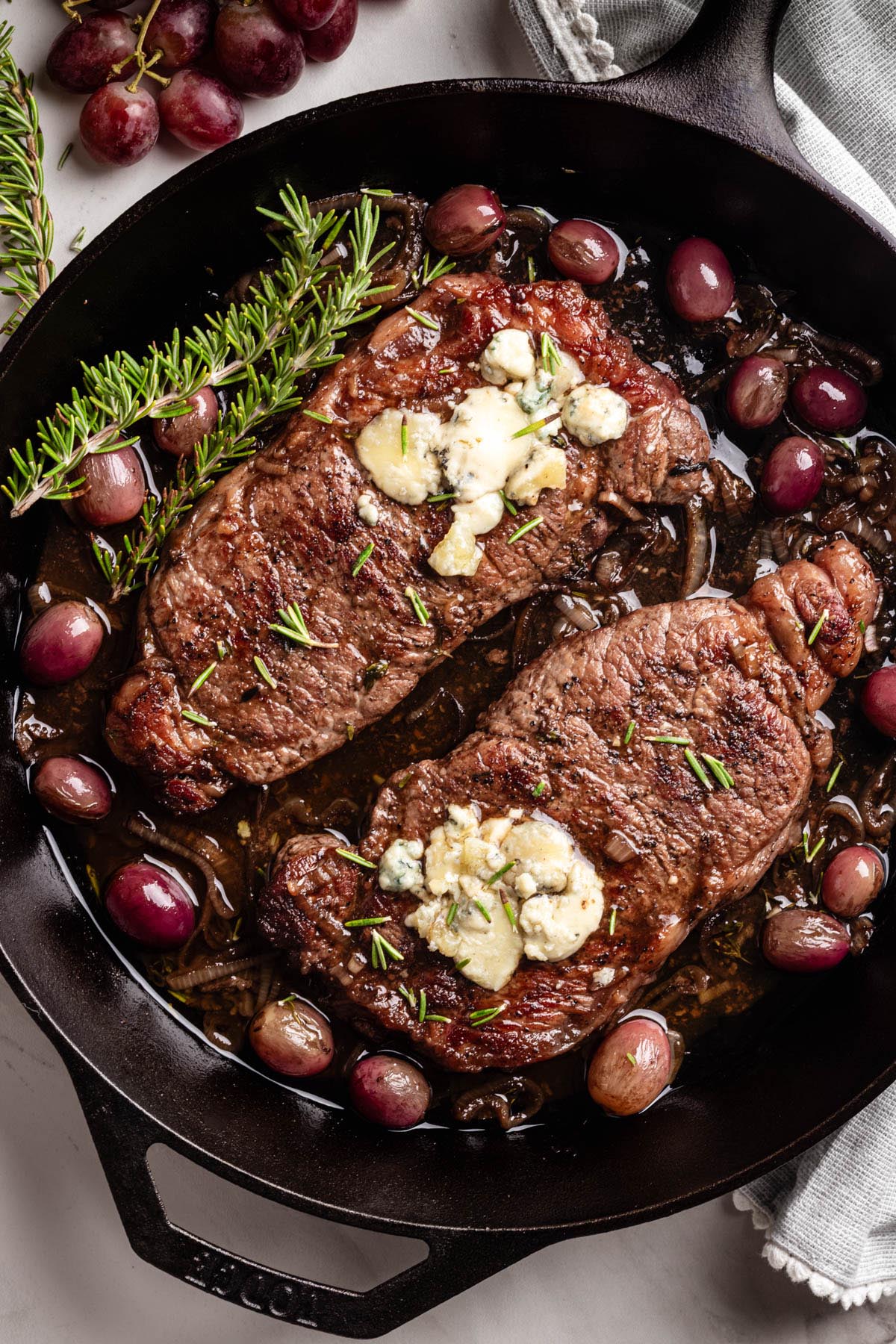 Close-up of seared strip steaks in a cast iron skillet, topped with melted blue cheese, garnished with fresh rosemary, and surrounded by roasted red grapes and caramelized shallots.