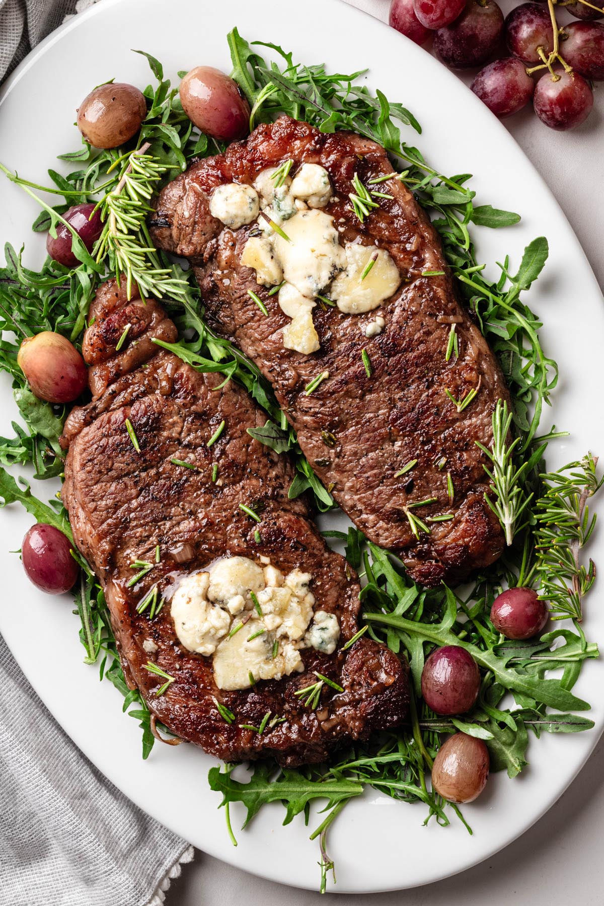 Two strip steaks topped with melted blue cheese, served on a white platter with arugula, roasted red grapes, and fresh rosemary sprigs.